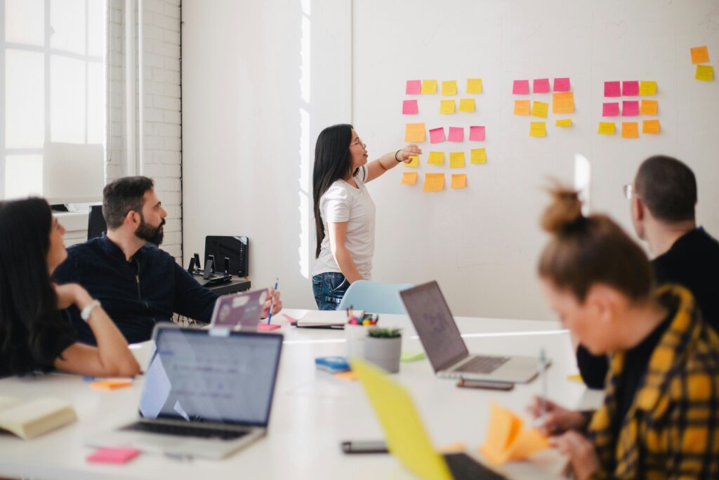 Kvinnelig kollega peker på en tavle med post-it lapper, mens andre kollegaer sitter rundt et bord og noterer og skriver på PC. Foto