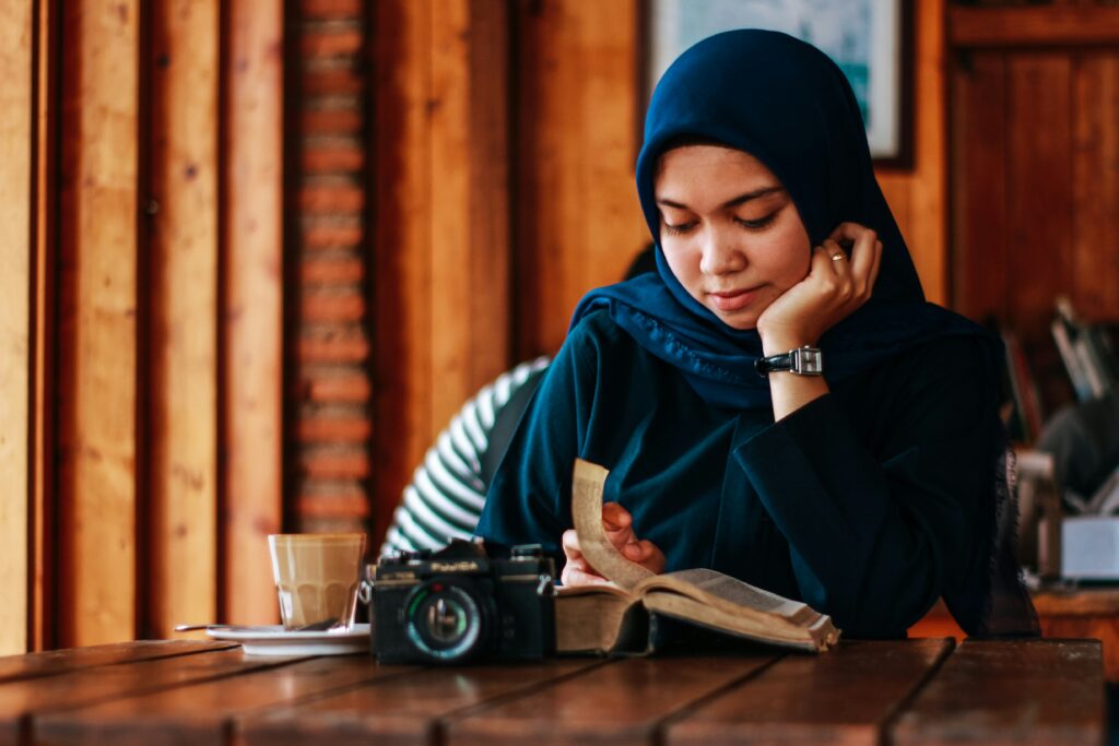 Kvinne med hijab sitter ved et bord og leser i en bok. Foto