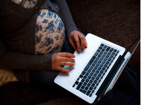 En gravid kvinne sitter med en bærbar PC i fanget, med hendene på tastaturet. Foto