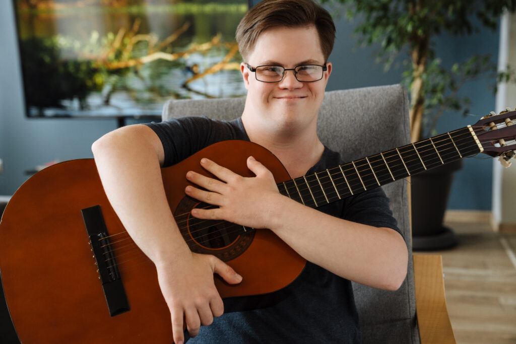 Ung mann med downs syndrom sitter på en sofa og holder rundt en gitar. Foto