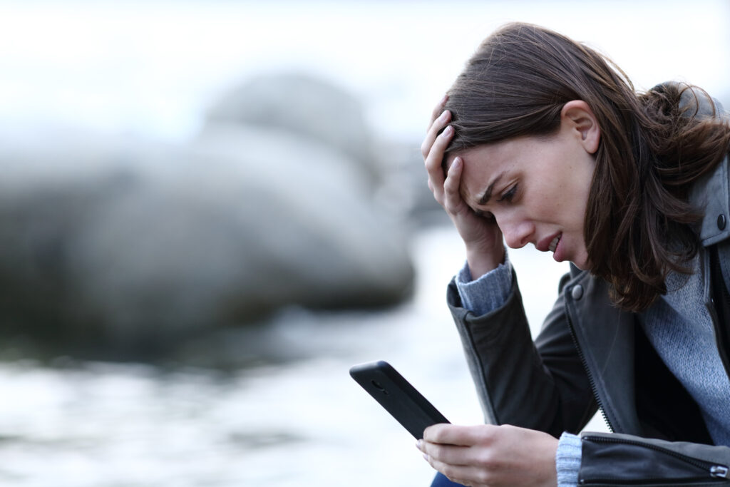 Bilde av fortvilet kvinne som sitter på en strand og holder seg i pannen mens hun ser på mobiltelefon. Foto