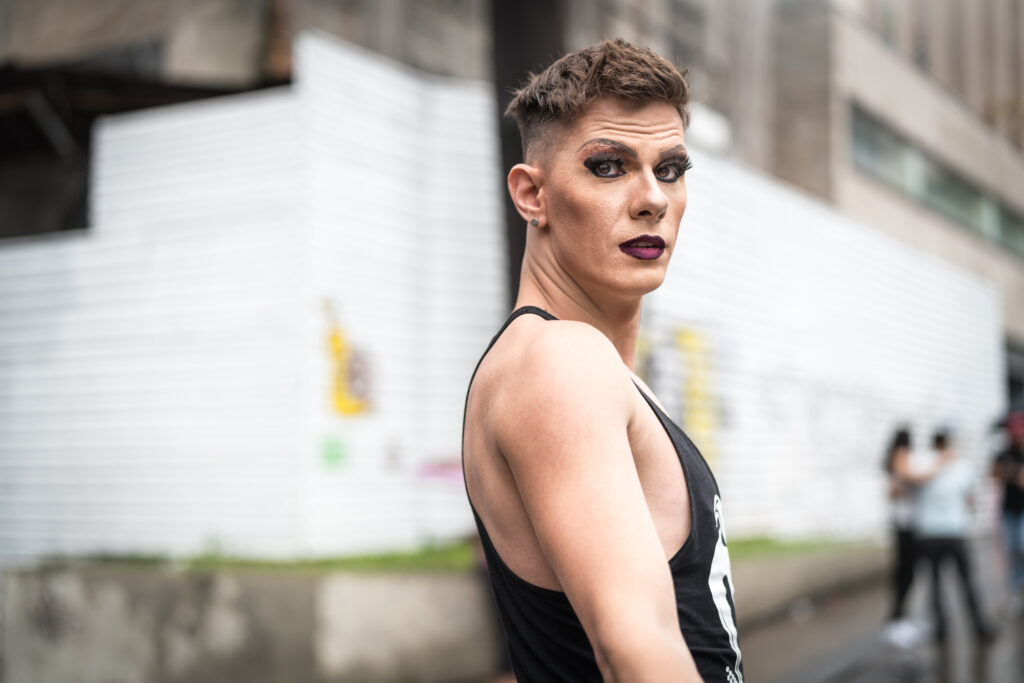 Mann med sminke og leppestift iført svart singlet poserer på et gatehjørne. Foto