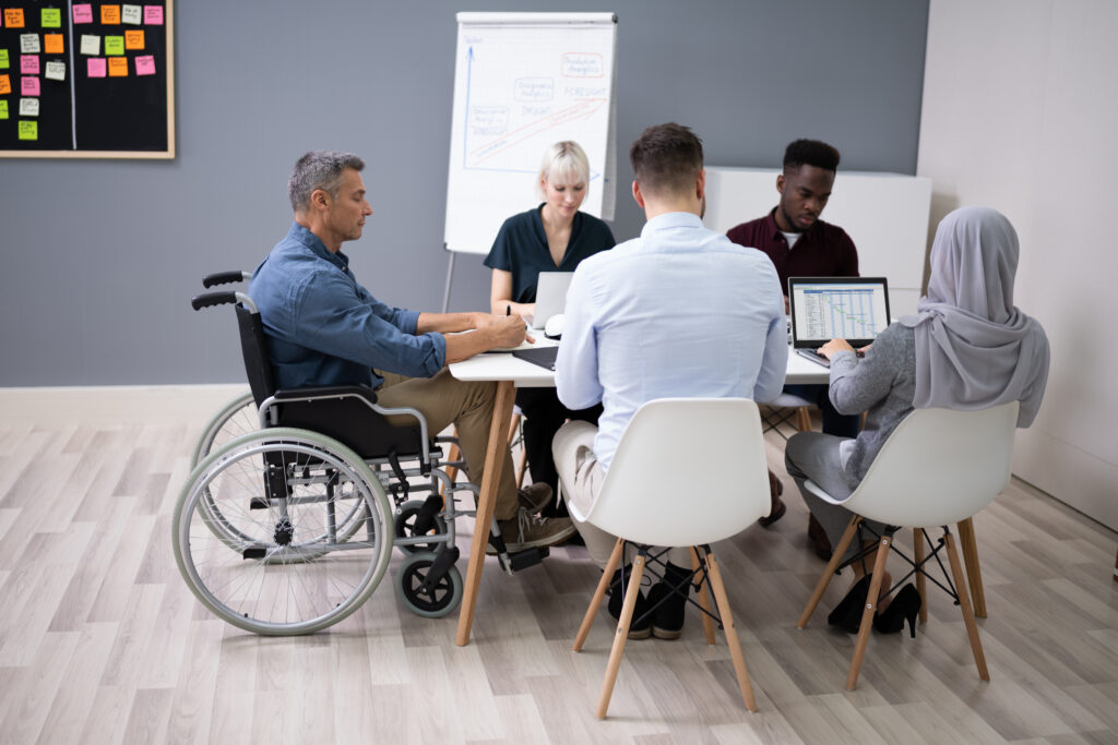 En gruppe på fem arbeidstakere sitter rundt et bord og diskuterer. En av dem sitter i rullestol. Foto