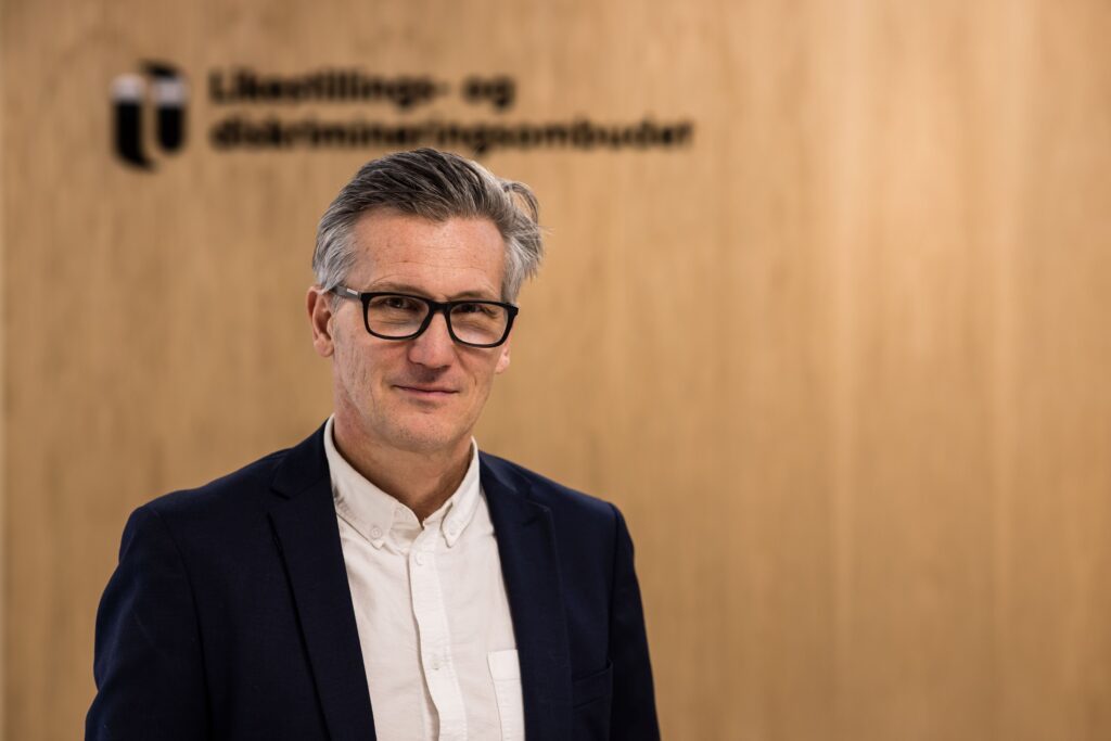 Bilde av en mann i hvit skjorte og mørk dressjakke, med sorte briller og grålig hår.