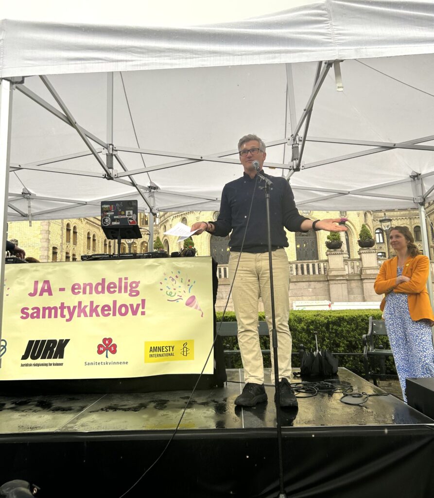 En mann med briller og et ark i hånden står på en scene foran Stortinget. Ved siden av han står teksten: JA - endelig samtykkelov!