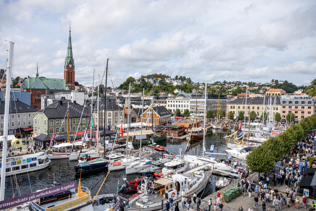Oversiktsbilde av Arendals sentrum under Arendalsuka, med seilbåter og trebåter ved kaia, folkemengde på bryggekanten og kirketårnet i bakgrunnen