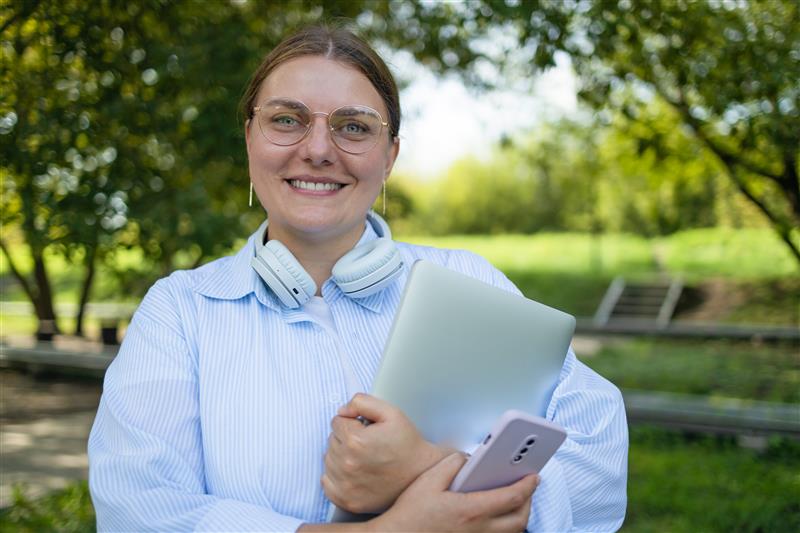 Jente med stripete blå skjorte og briller står med høretelefoner rundt nakken, og PC og mobil i hånda.