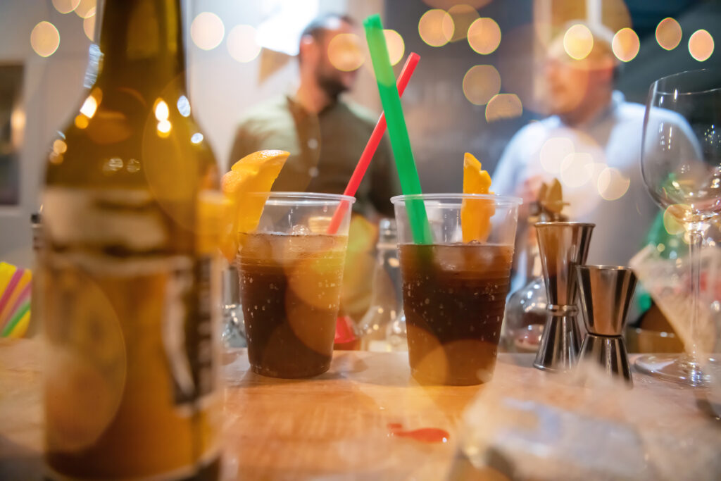 Bildet viser drinker med sugerør på et bord i tillegg til ølflasker og shot-glass. I bakgrunnen ses mennesker som snakker med hverandre. Foto