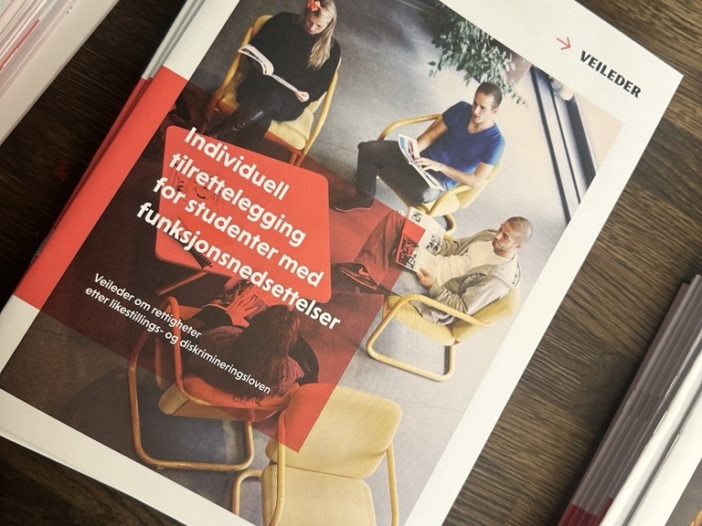 Rapport i A4-format ligger på et bord. Rapportens forside viser bilde at fire studenter som sitter rundt et bord. Tittelen på rapporten er "Individuell tilrettelegging for studenter med funksjonsnedsettelser. Foto