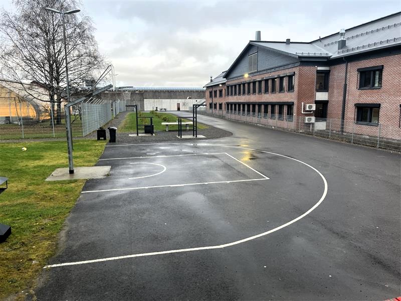 Bredtvet fengsel sin bakgård med mursteinbygg i bakgrunnen og basketballbane i forgrunnen omringet av gjerde. Foto