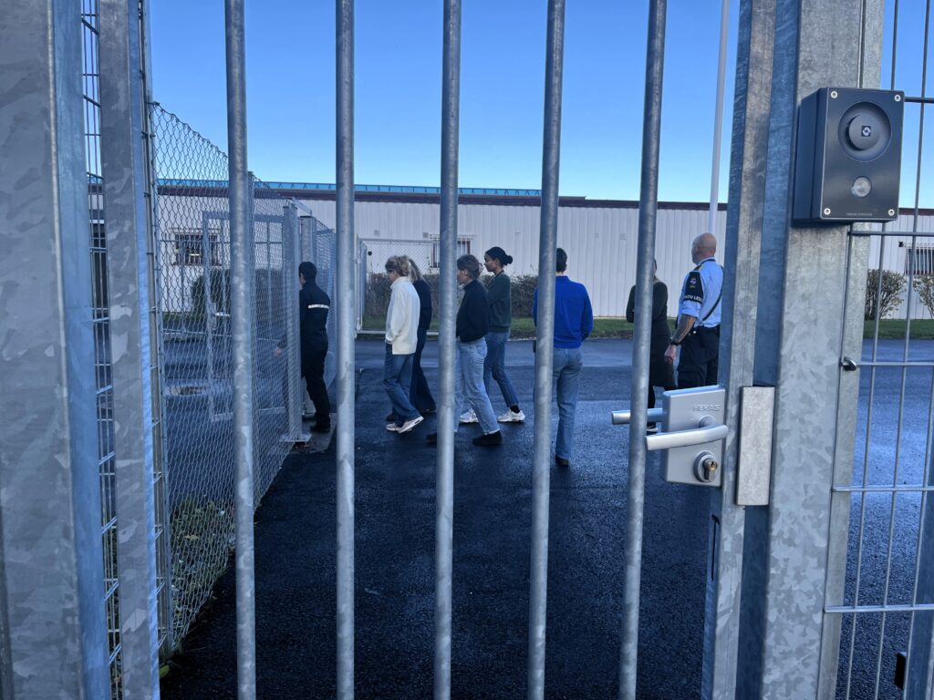 Bredtvet fengsel sin adgangsport med gittegjerde ses i forgrunnen mens folk spaserer inn porten i bakgrunnen. Foto