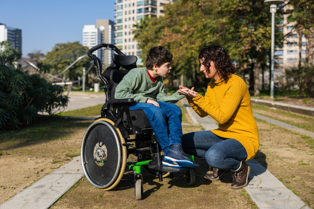 En kvinne sitter på huk og holder hånden til et barn med funksjonsnedsettelse som sitter i rullestol. Foto