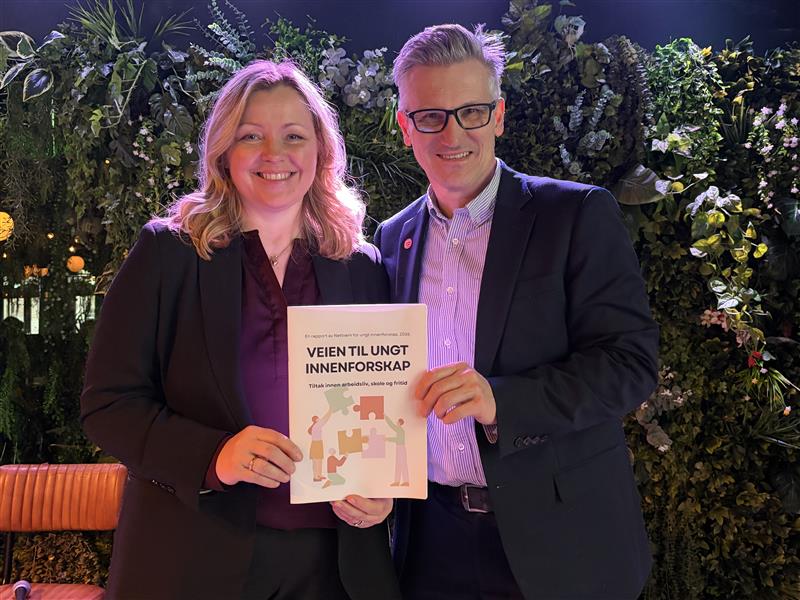 En kvinne og en mann holder rapporten Veien til ungt innenforskap mellom seg. Begge er kledd i mørk blazer.