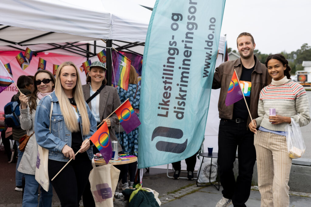 Fem mennesker står samlet ved en stand. Flere av dem holder pride-flagg i hendene. I midten av bildet ser du en lyseblått beach-flagg med logoen til Likestillings- og diskrimineringsombudet.