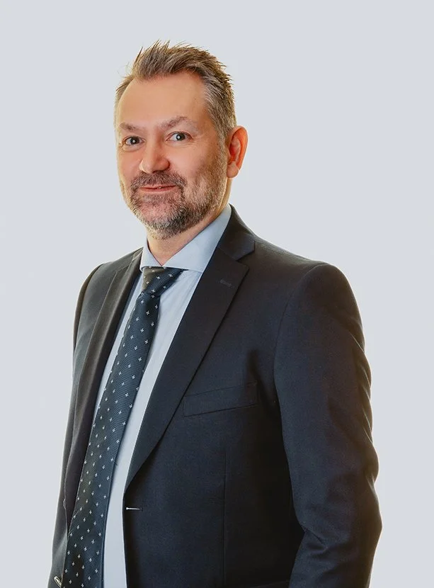 Bildet er av en mann med kort, brunt hår som ser inn i kameraet. Han har på seg dress og mønstrete slips, med blå skjorte inni. Bildet har grå bakgrunn. Mannen Bertil Christian Hast Rønnestad, advokat.