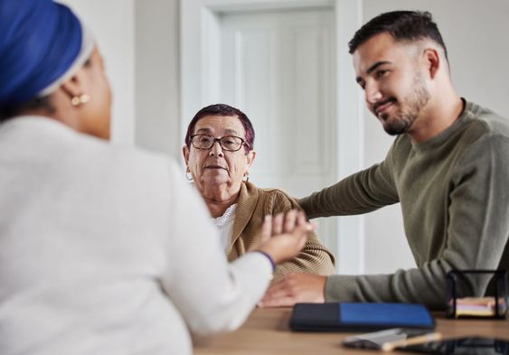 To kvinner og en mann sitter rundt et bord på et kontor. En av kvinnene gir de to andre råd. Hun har på seg hvit overdel og blått hodeplagg. Den andre kvinnen, som mottar råd, har på seg beige overdel. Hun har kort hår, briller, og er eldre enn de to andre på bildet. Den tredje personen i bildet har sort hår og sort skjegg. Overdelen hans er grå. Han holder armen rundt den eldre kvinnen.