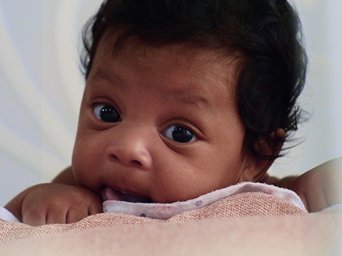 baby med svart hår og brunlig hud ser inn i kamera med nusselig uttrykk